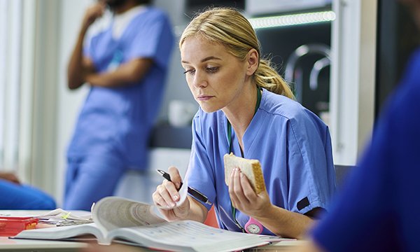 Nutrition for nurses on shift: a nurse sits at a table in a break room holding a sandwich in one hand while leafing through a document