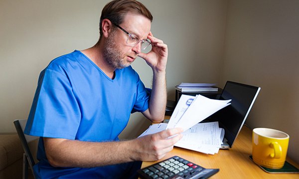 Nurses’ pay and pension worries: concerned nurse sits at desk, head in hand as he read documents that could be bills or bank statements