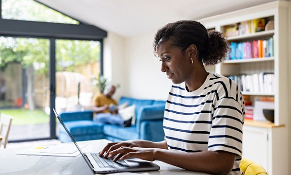 Written reflective account: a woman sits at a table in her living room typing on a laptop