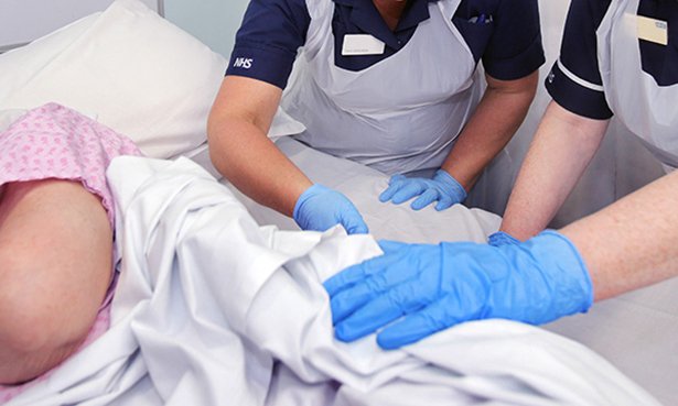 Wound management: two hospital nurses appear to attend to a patient’s sacrum, which is protected from view by a sheet