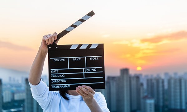A person holds a clapper board on a film set, standing at the top of a building overlooking a city skyline
