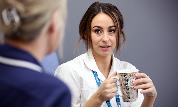 nurse looks apprehensive, talking to a colleague, mug in hand